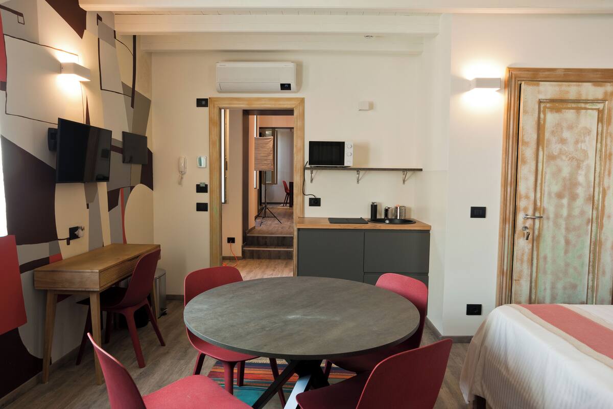 Quarto de hotel compacto com uma mesa redonda, cadeiras vermelhas, cama, cozinha compacta e televisão fixada na parede.