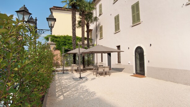 A courtyard with outdoor seating, palm trees, and a building with green shutters. - ANNAMA Coliving (Brescia)