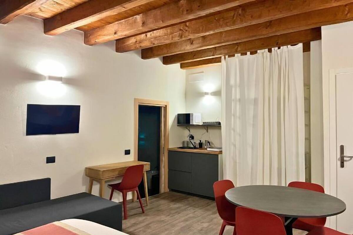 Quarto compacto com cama, escrivaninha, pequena cozinha e mesa de jantar com cadeiras.