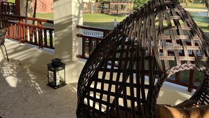 A wicker hanging chair with a cushion on a covered porch with a view of a building and palm trees. - Magic Arena Blanca Beach Suite (Punta Cana)