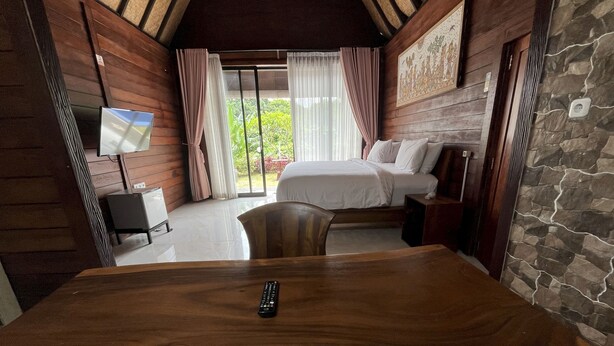 A wooden dining table with a remote control, a bed with white linens, a TV on the wall, and a stone wall. - Belvilla Darshana Bali Zoo (Batubulan)