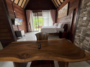 A wooden dining table with a remote control, a bed with white linens, a TV on the wall, and a stone wall. - Belvilla Darshana Bali Zoo (Batubulan)