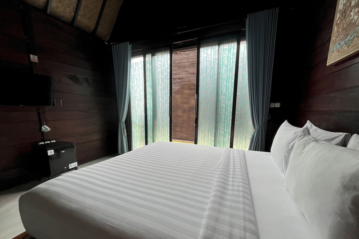 A bedroom with a large bed, a wooden headboard, a nightstand, a TV, and a window with curtains.