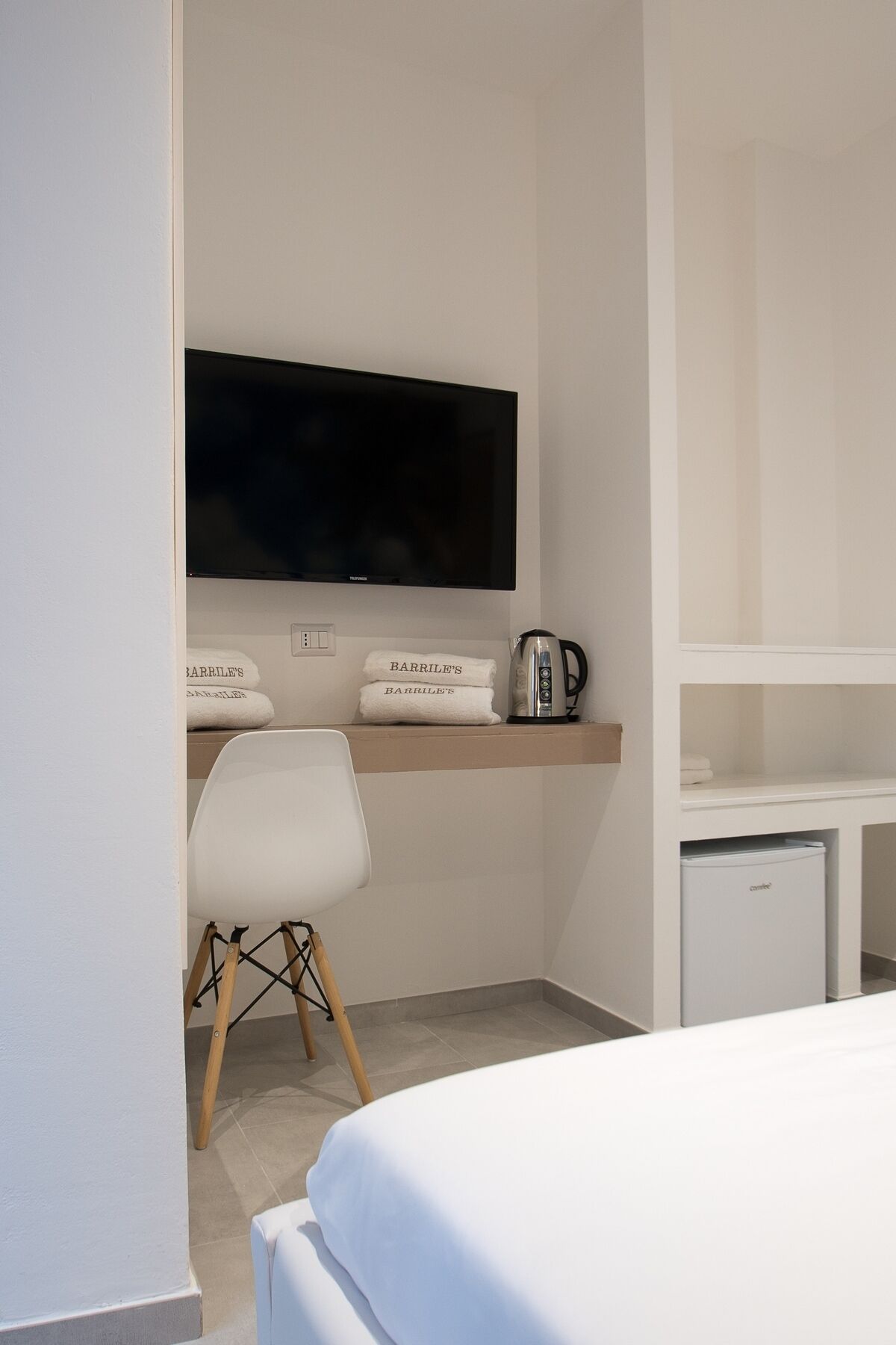 Ett hotellrum med en säng, ett skrivbord med en platt-TV, en stol, en hylla med handdukar och en vattenkokare.