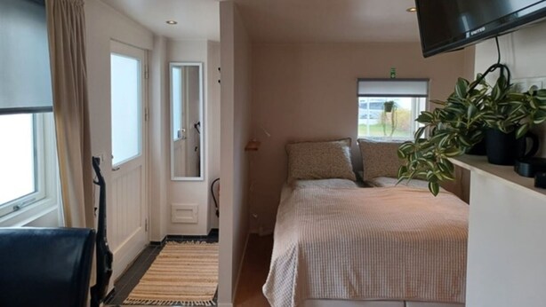 A bedroom with a bed, a TV mounted on the wall, a window with a view, and a door leading to another room. - Nordurgata 43 (Akureyri)