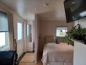 Un dormitorio con cama, televisor montado en la pared, ventana con vistas y puerta que da a otra habitación.