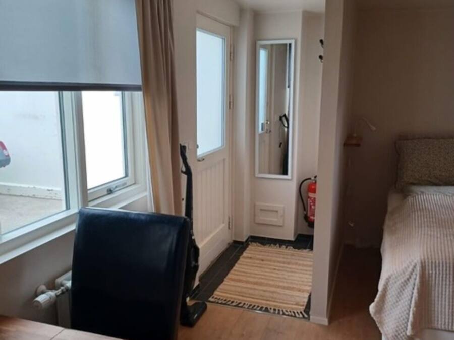 A hotel room with a wooden floor, a round table, a bed with a striped blanket, a window with curtains, and a mirror on the wall.