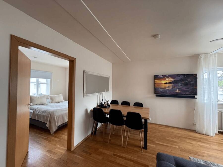 A modern room with a wooden floor, a bed, a dining table with chairs, a TV, and a large window with curtains.