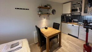 A compact kitchen with white cabinets, a microwave, and a dishwasher. There is a wooden dining table with chairs, a small shelf with plants, and a radiator. - Nordurgata 43 (Akureyri)