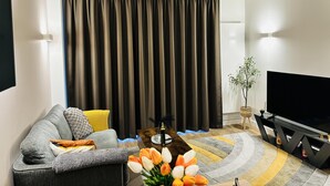 A modern living room with a grey sofa, a flat-screen TV, a coffee table, and a large window with dark curtains. - Your London Escape Luxurious 1 Bedroom Apartment (Wembley)