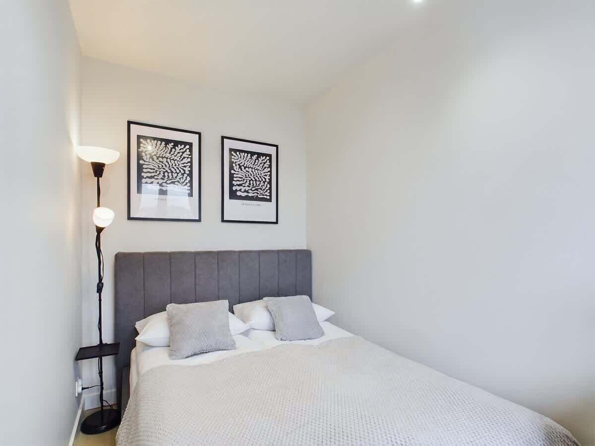 Ein ordentlich eingerichtetes Schlafzimmer mit einem Bett, Kissen, einer Nachttischlampe und zwei gerahmten Kunstwerken an der Wand.