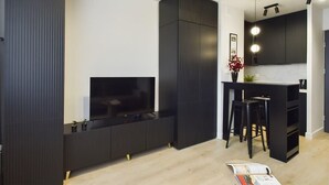 A modern kitchen with dark cabinetry, a built-in TV, and a bar area with stools. - Modern Studio with Workspace | Close to Metro (Warszawa)