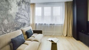 A living room with a beige sofa, a small coffee table, and a large window with curtains. - Modern Studio with Workspace | Close to Metro (Warszawa)