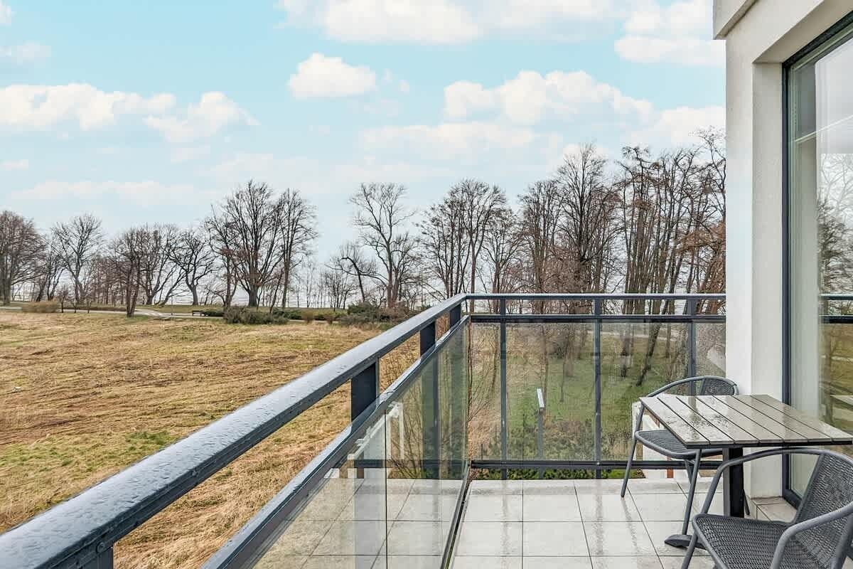 Un balcon doté d’une balustrade en verre, d’une table et de chaises, offrant une vue sur un champ verdoyant et des arbres.