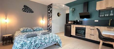 A compact bedroom with a bed, bedside lamps, and a small desk with a chair. Adjacent is a kitchenette with a microwave, coffee maker, and cabinets.