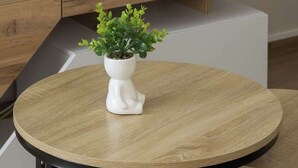 A round wooden table with a white vase containing green plants, positioned on a wooden floor with a black metal frame.