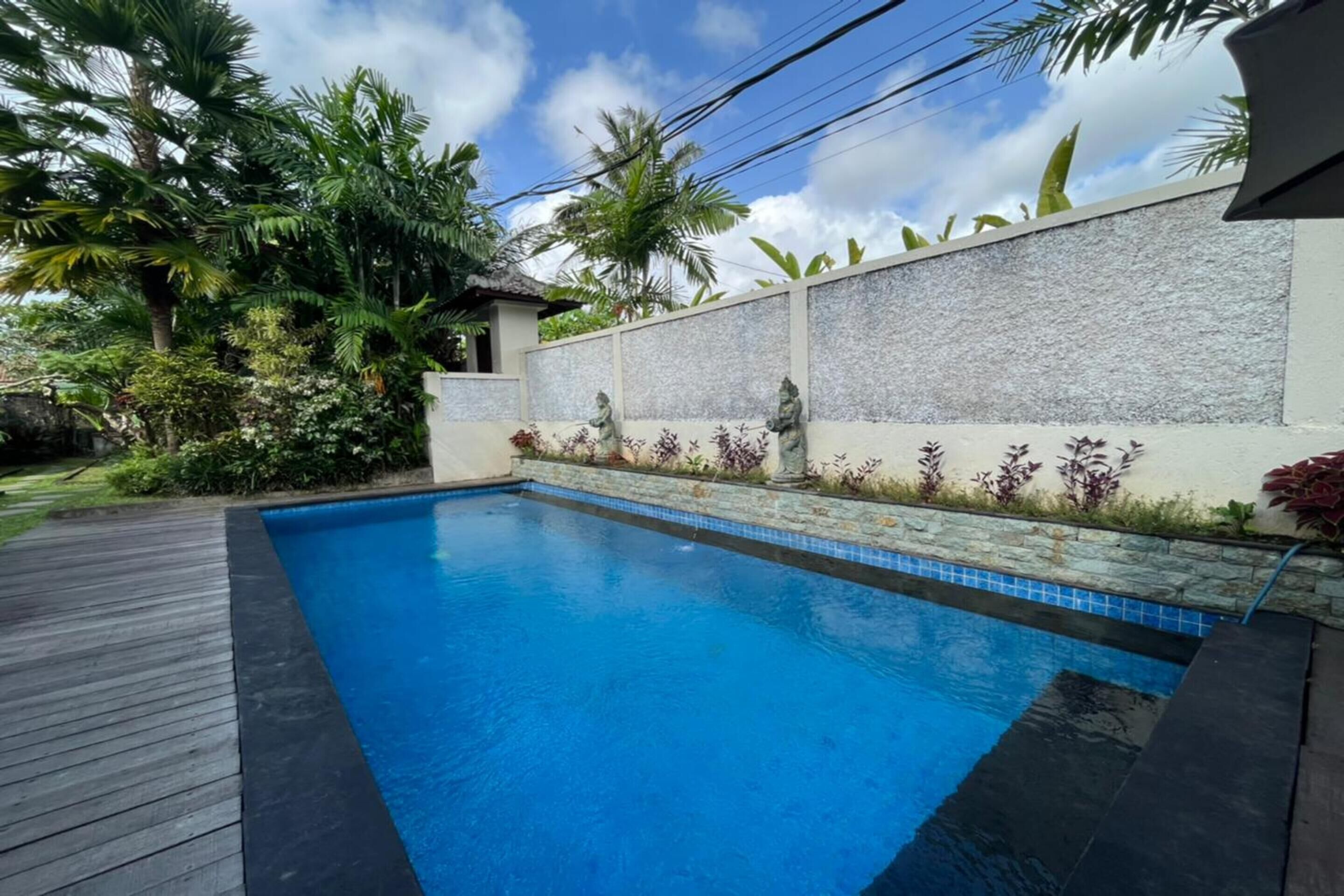 Una piscina con cubierta de madera, rodeada de plantas tropicales y un muro de piedra con estatuas.