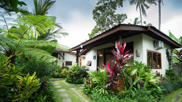 A bungalow with a thatched roof, surrounded by lush tropical vegetation and a stone pathway. - Belvilla Chaitanya Niketan (Ubud)
