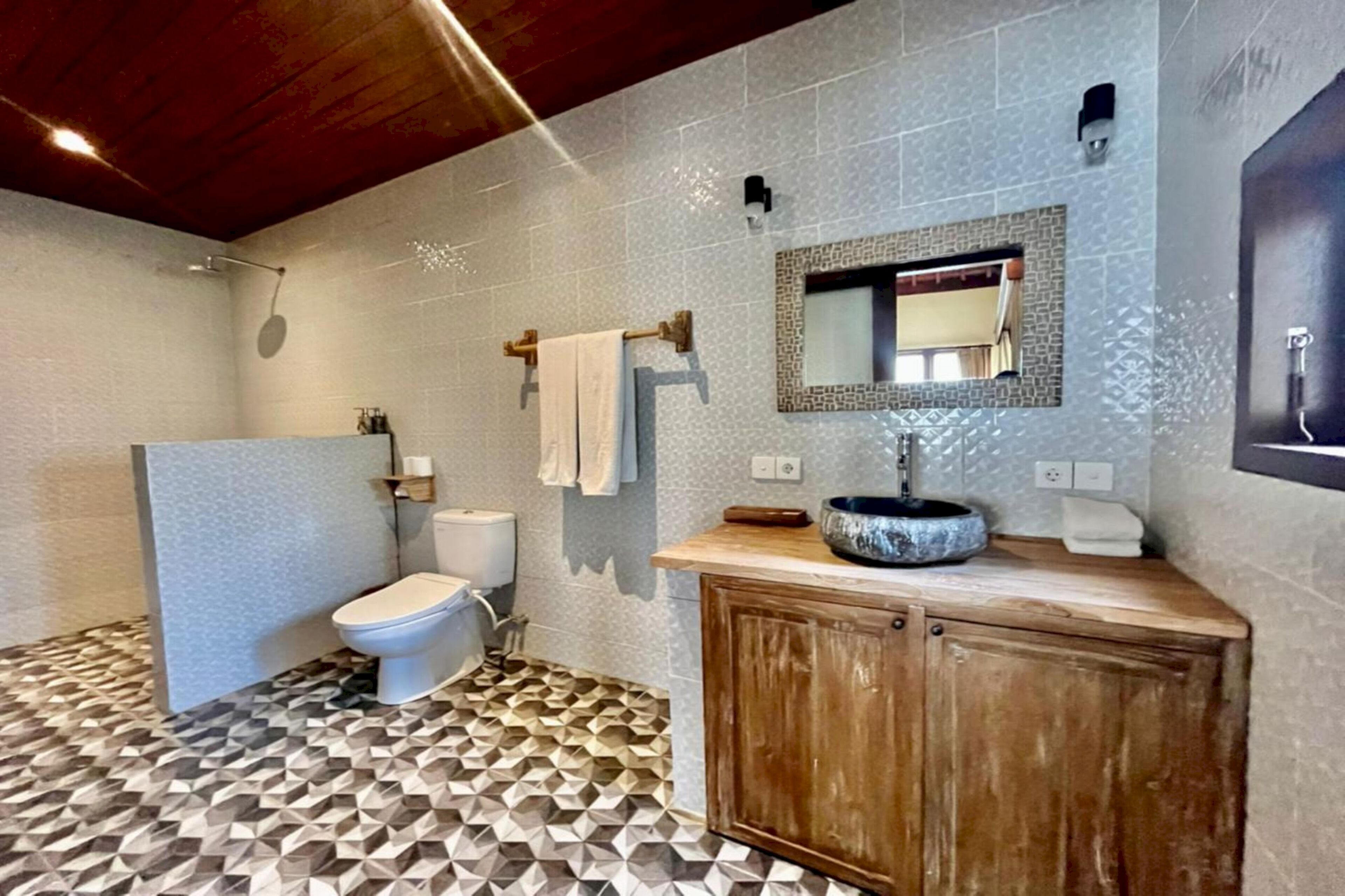 Un baño con piso de patrón geométrico, tocador de madera, lavamanos, inodoro y zona de ducha.