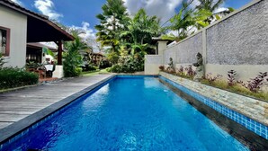 A swimming pool with a wooden deck, surrounded by lush greenery and a stone wall. - Belvilla Chaitanya Niketan (Ubud)