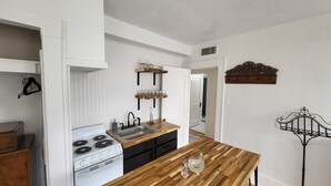 A kitchen with white cabinets, a wooden countertop, a stove, and a sink. There are shelves above the counter holding glasses and a decorative metal piece on the wall.