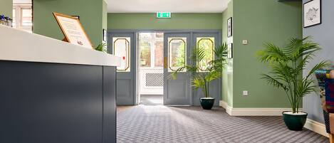 Uma área de recepção com balcão de check-in, parede verde, carpete estampado e plantas em vasos.