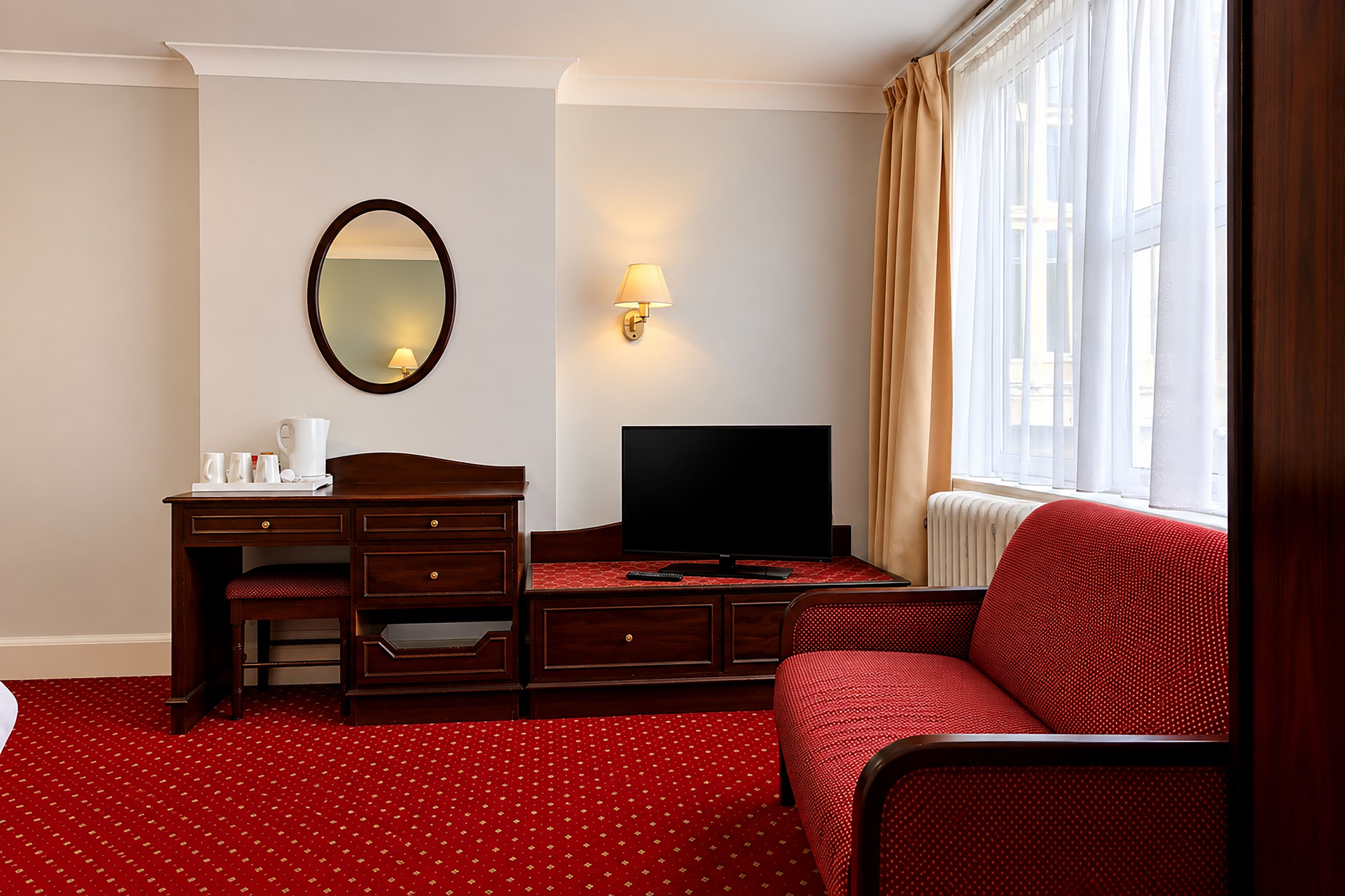 Habitación de hotel con escritorio de madera, televisor, espejo, sofá rojo estampado y ventana con cortinas.