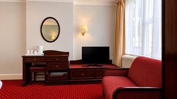 Habitación de hotel con escritorio de madera, televisor, espejo, sofá rojo estampado y ventana con cortinas.
