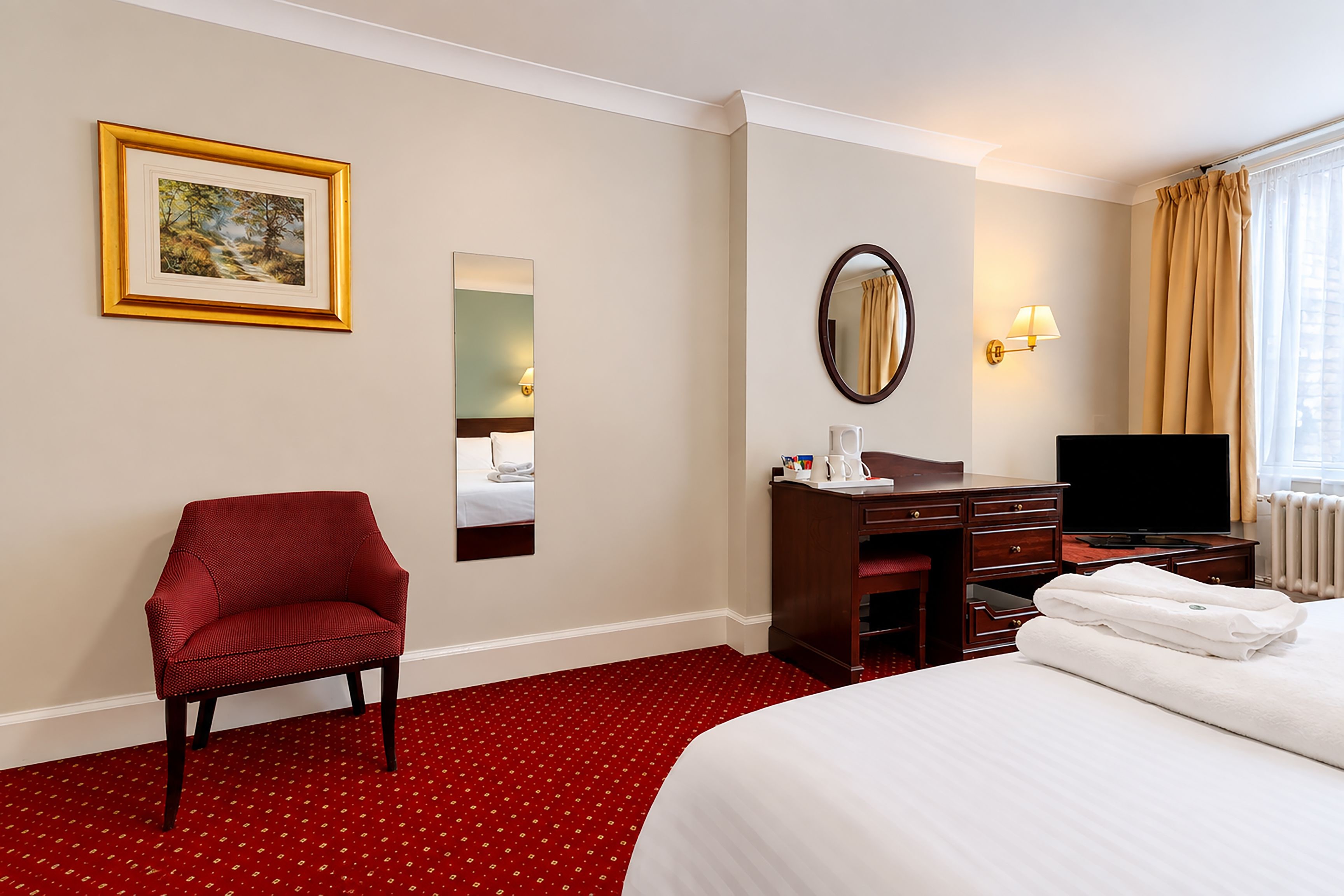 Une chambre d’hôtel comprenant un lit, un bureau, une chaise, une télévision, un miroir, un tableau et une fenêtre avec des rideaux.