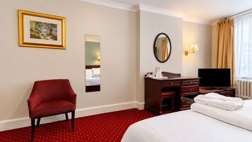 Une chambre d’hôtel comprenant un lit, un bureau, une chaise, une télévision, un miroir, un tableau et une fenêtre avec des rideaux.