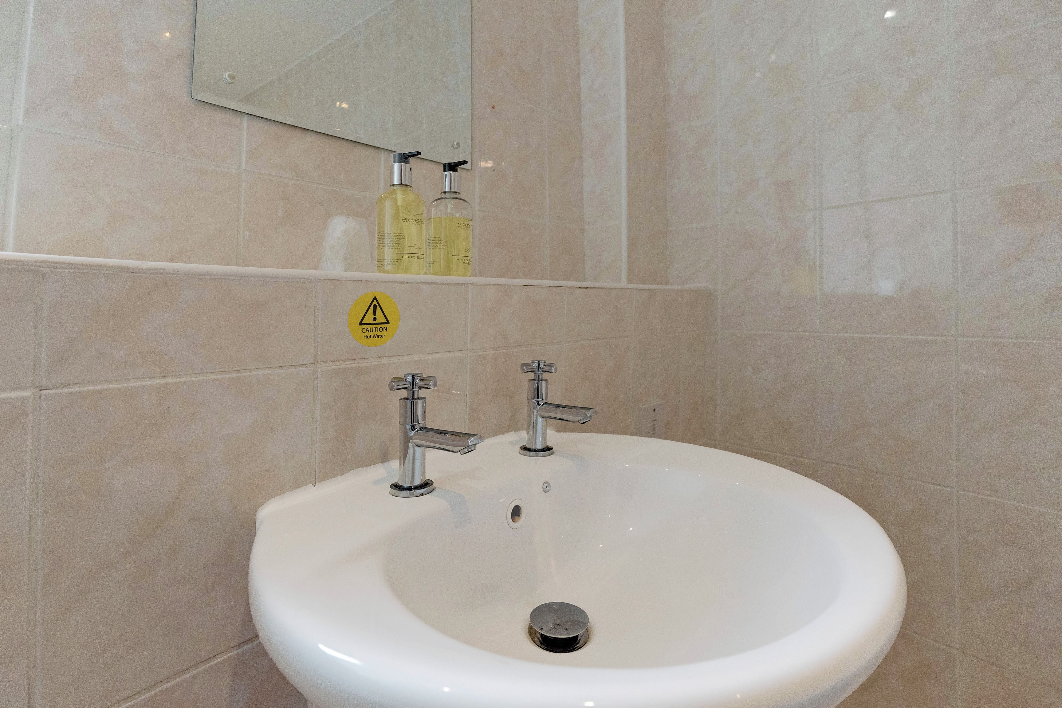A bathroom with a white sink, two faucets, and a mirror above. There is a caution sign for hot water on the wall.