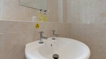 A bathroom with a white sink, two faucets, and a mirror above. There is a caution sign for hot water on the wall.