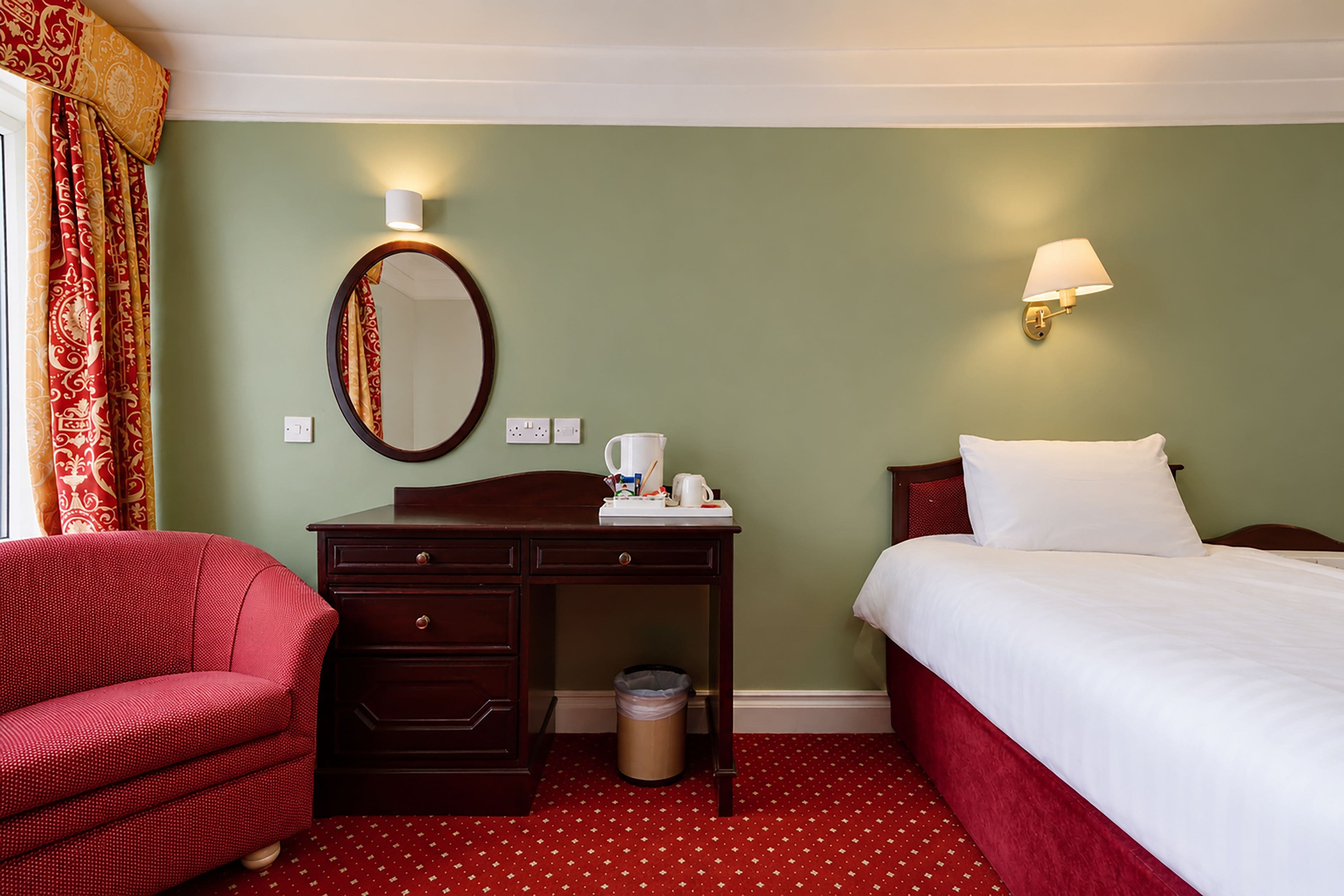 A hotel room with a bed, a desk with a mirror, a chair, and a nightstand.