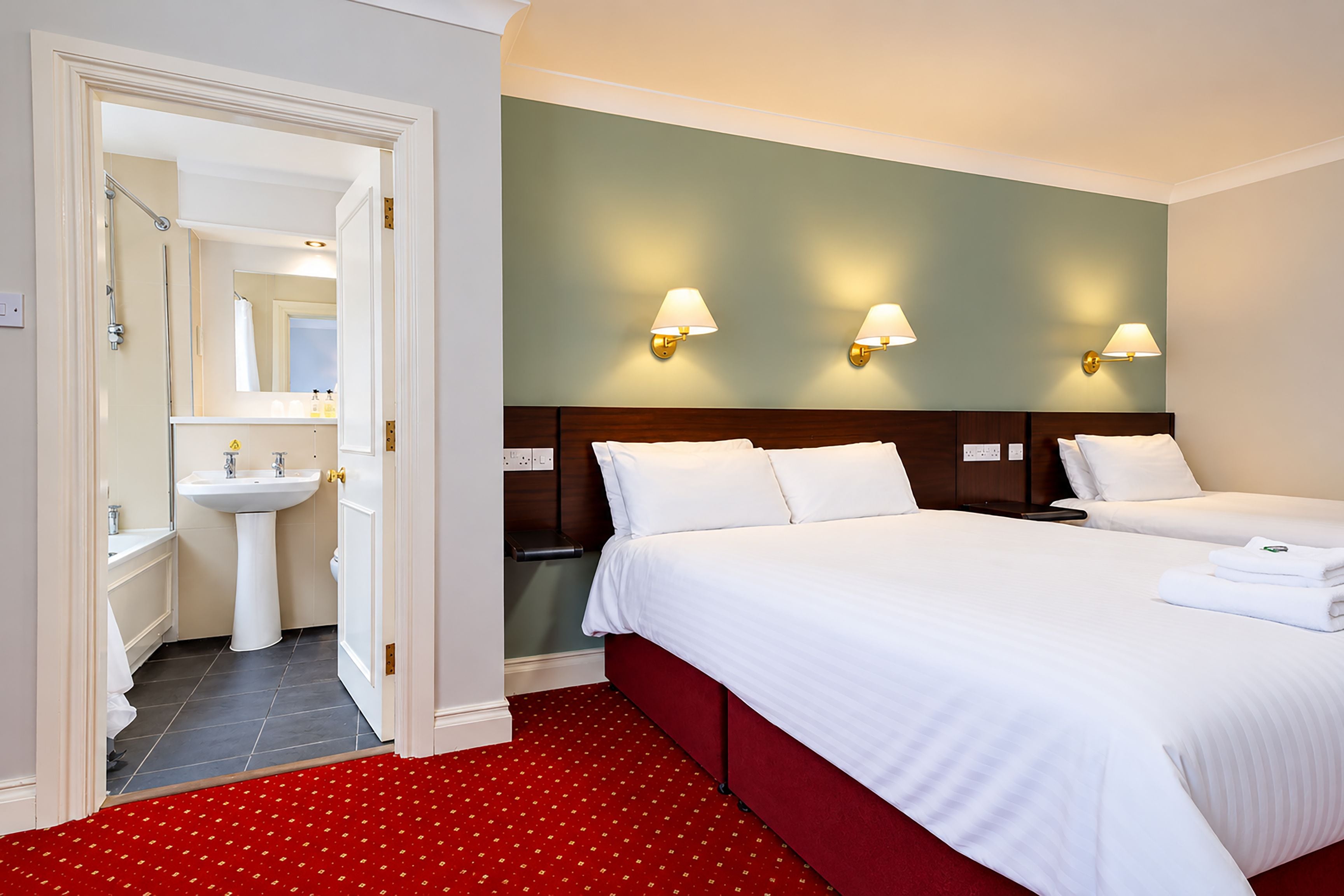 Une chambre d’hôtel comprenant un lit, deux tables de chevet, une commode, une moquette rouge, une salle de bain avec un lavabo et un miroir, et des lampes fixées au mur.