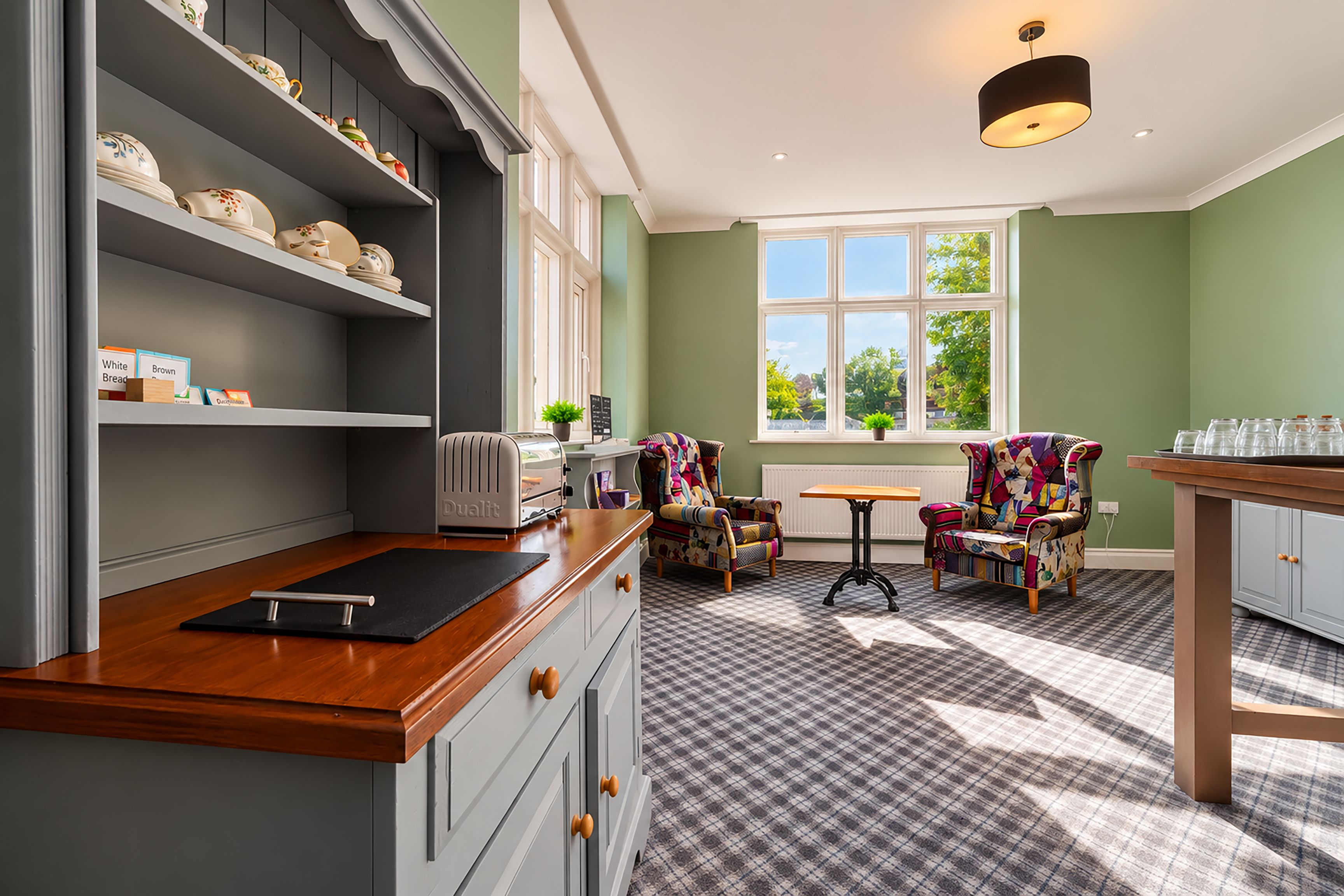 A kitchen with a wooden countertop, a toaster, and a chair with a colorful quilt. There is a window with a view of trees and a radiator on the wall.