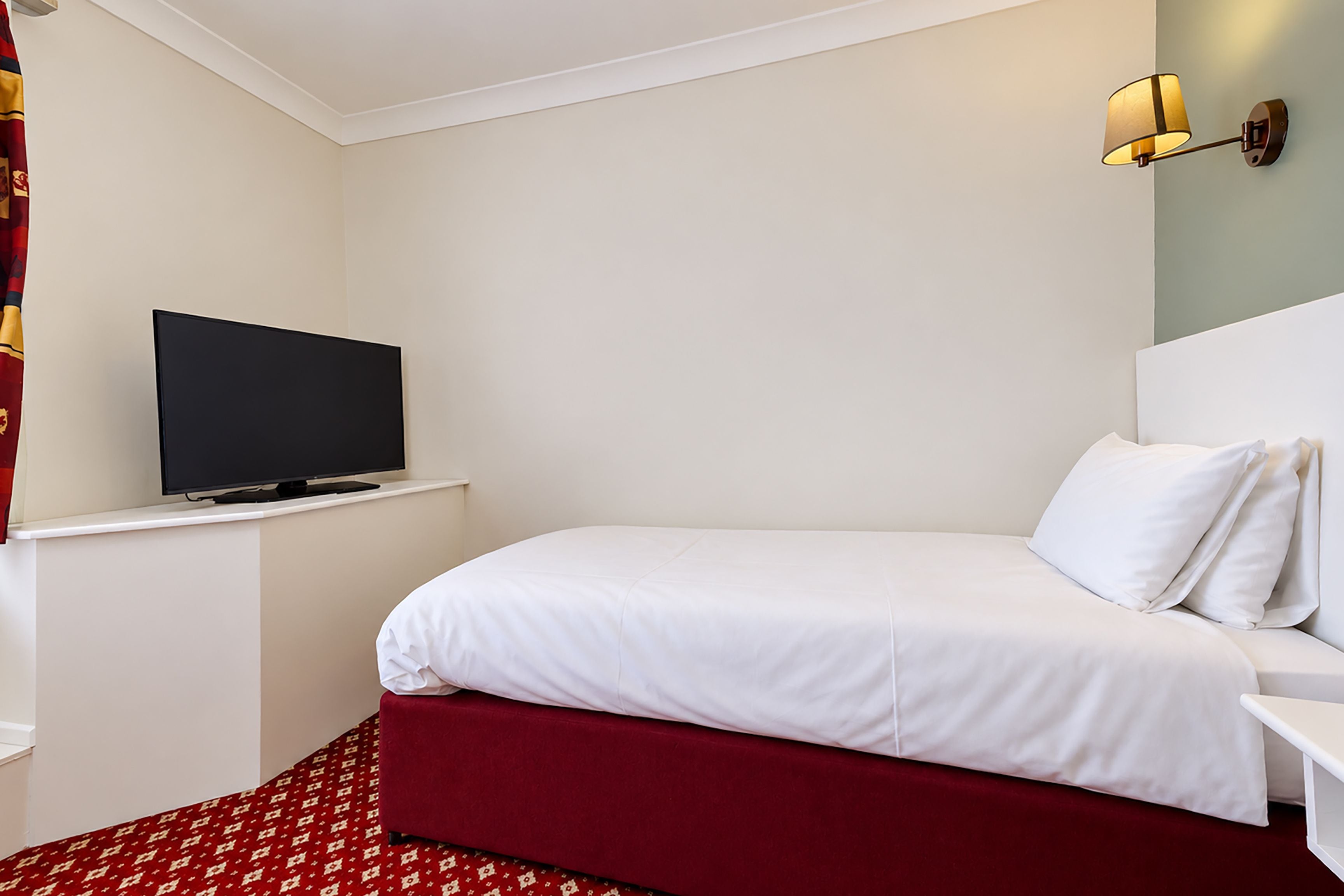 Ein Hotelzimmer mit Bett, Fernseher auf einem Schrank, einem roten Teppich und einer Wandleuchte.
