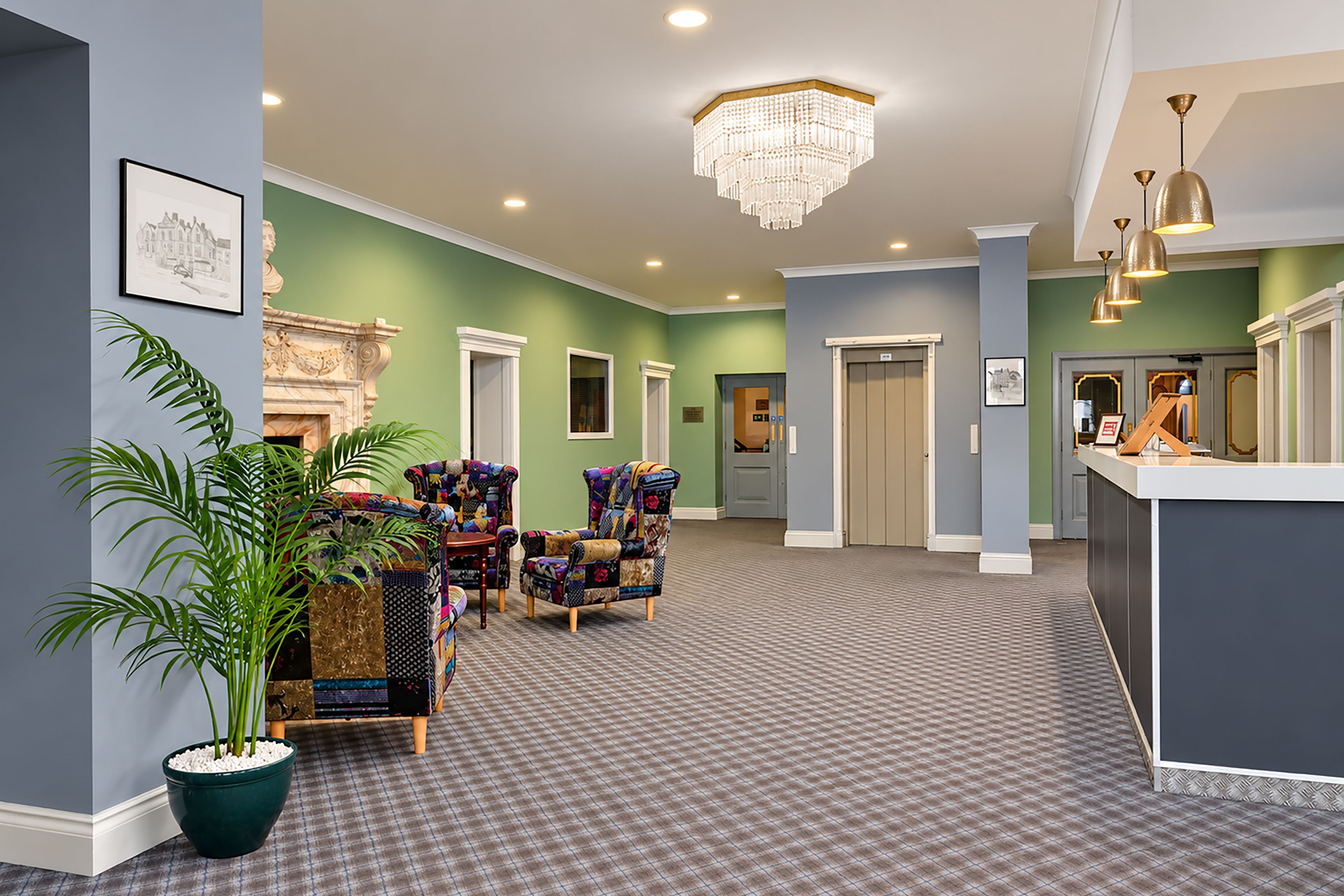 A hotel lobby with a check-in counter, seating area with patterned chairs, a large chandelier, and a potted plant.