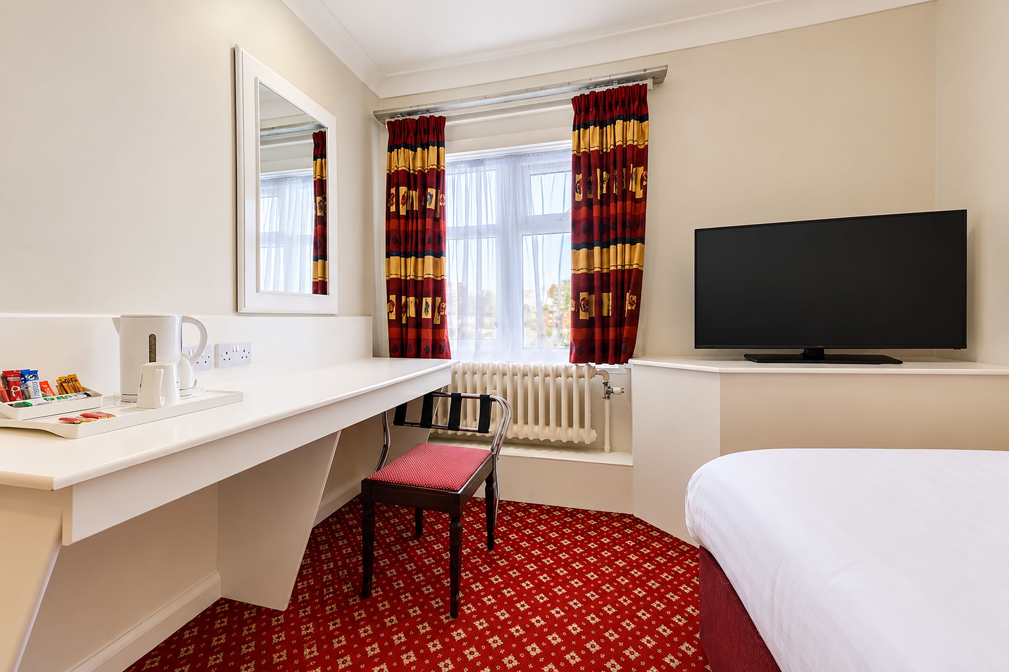 Habitación de hotel con escritorio blanco, televisor, alfombra roja estampada y ventana con cortinas.