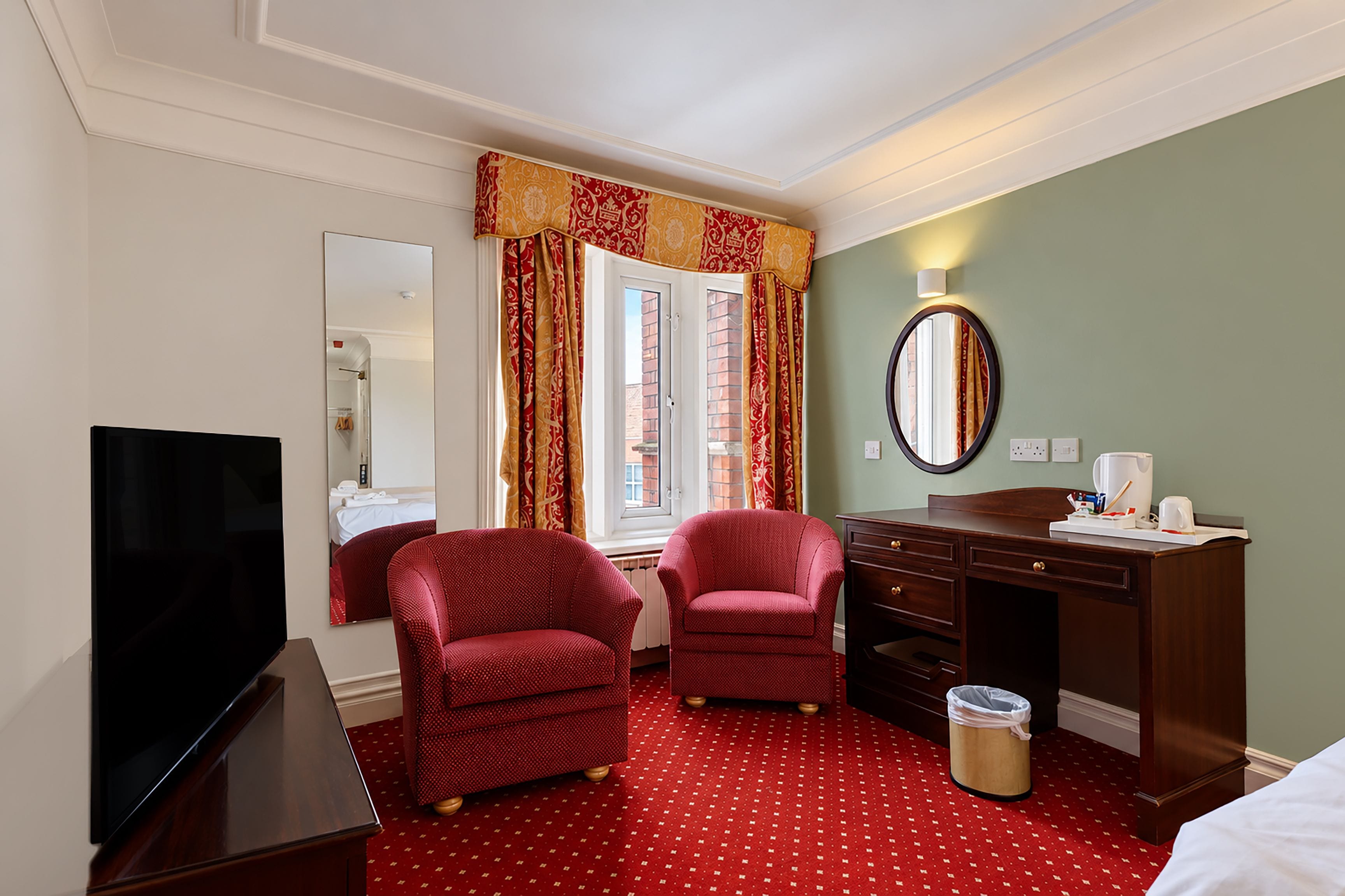 Een hotelkamer met een televisie, twee rode fauteuils, een bureau met een spiegel en een vloer met een rood tapijt.