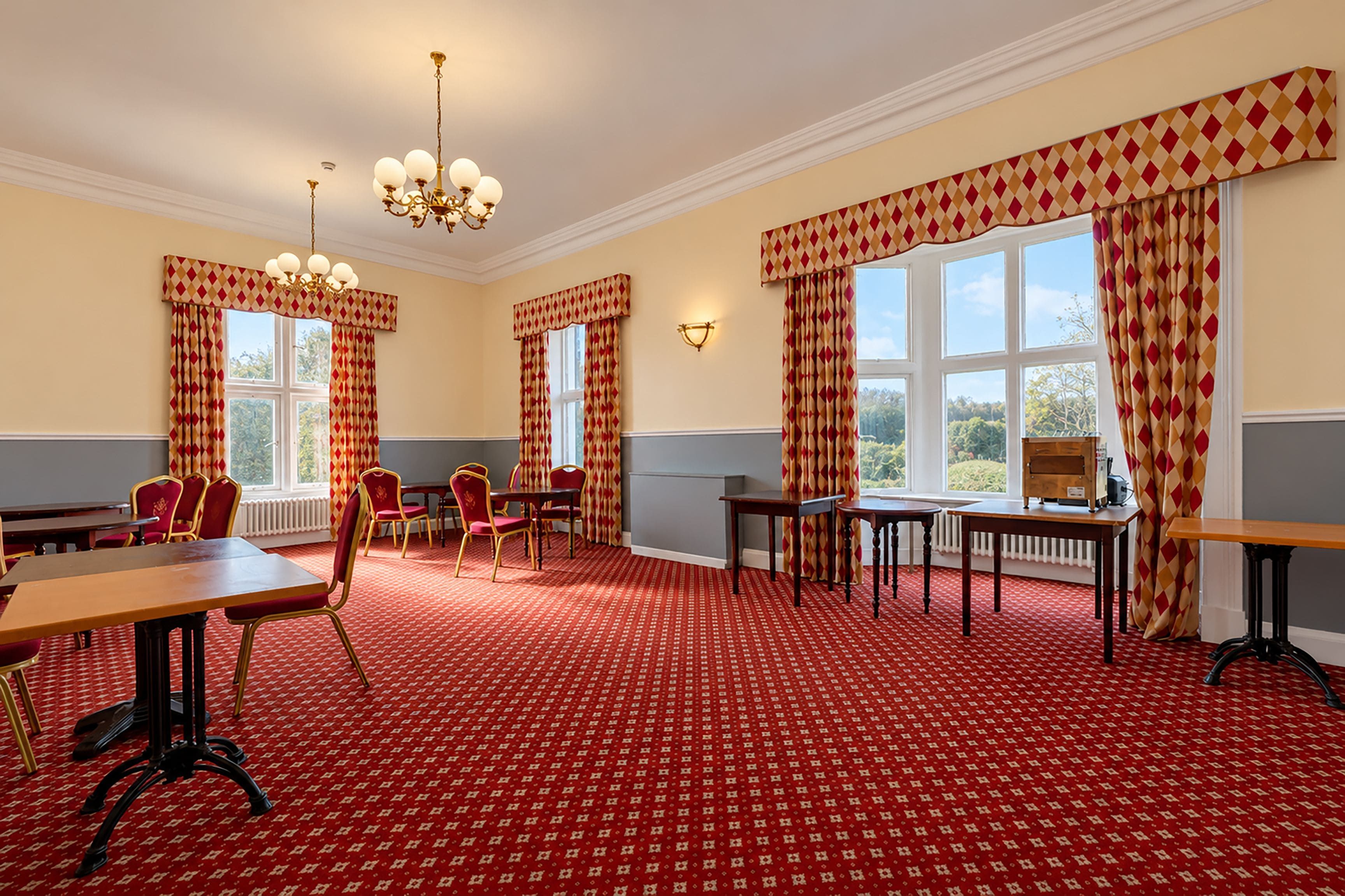 Ein Zimmer mit einem roten Teppich mit Muster, mehreren Tischen und Stühlen, großen Fenstern mit Vorhängen und einer Kristallleuchter.