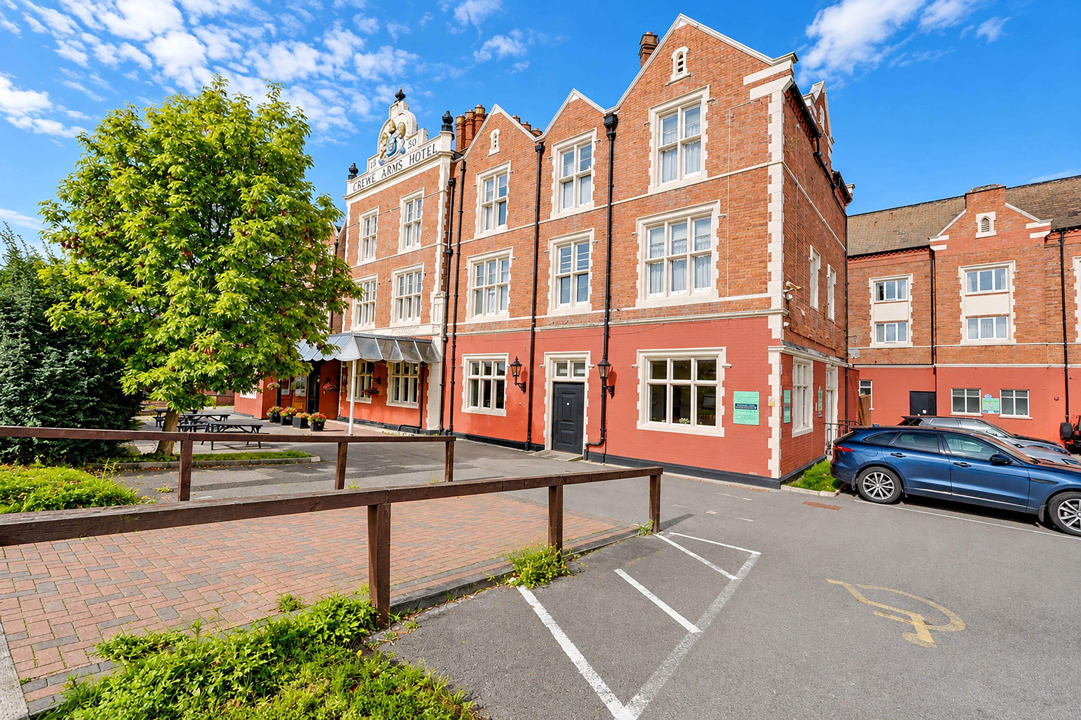 A red-brick building with a sign reading 'CRICKET ARMS HOTEL' and a parking area with a blue car.