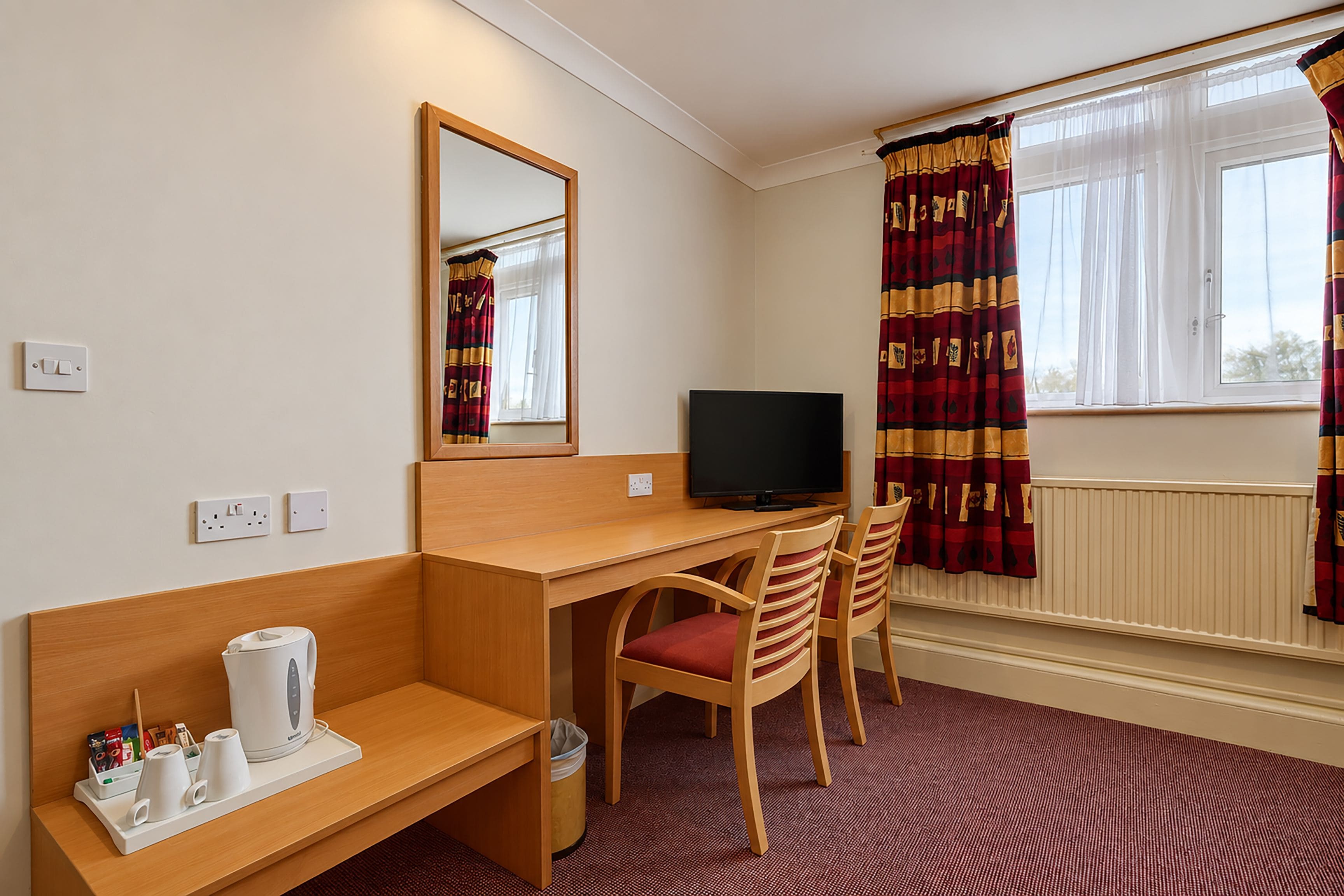 Une chambre d’hôtel avec un bureau en bois, une télévision, deux chaises, un miroir, une fenêtre avec des rideaux et un radiateur.