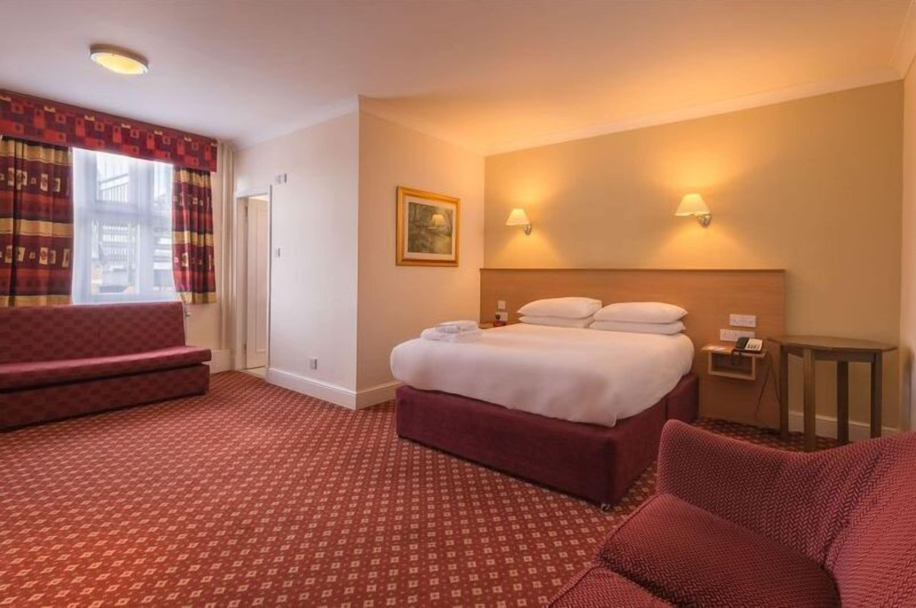A hotel room with a bed, a desk, a chair, a sofa, and a window with curtains.