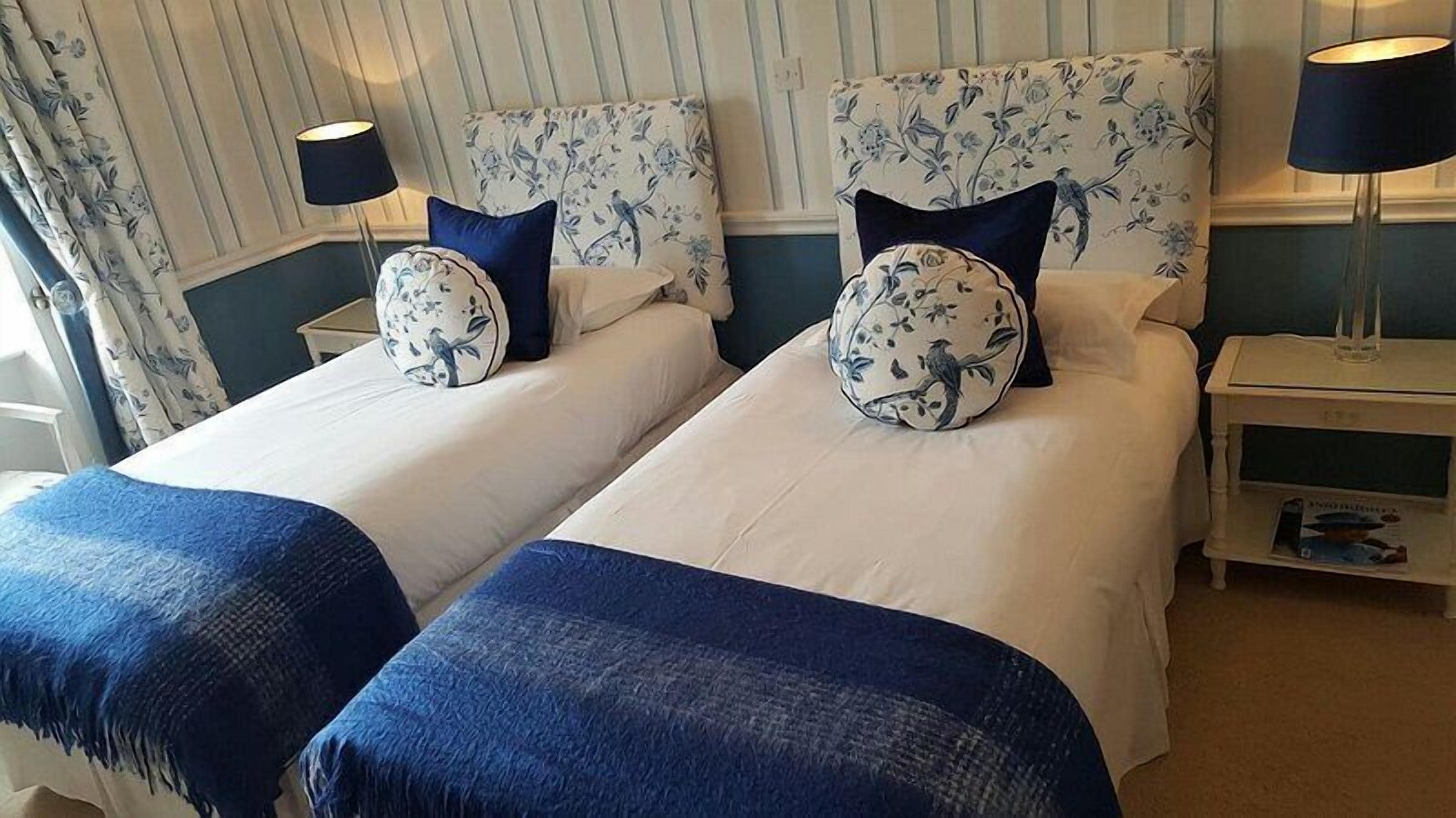 A hotel room with two single beds, each with a matching pillow and a blue throw blanket.