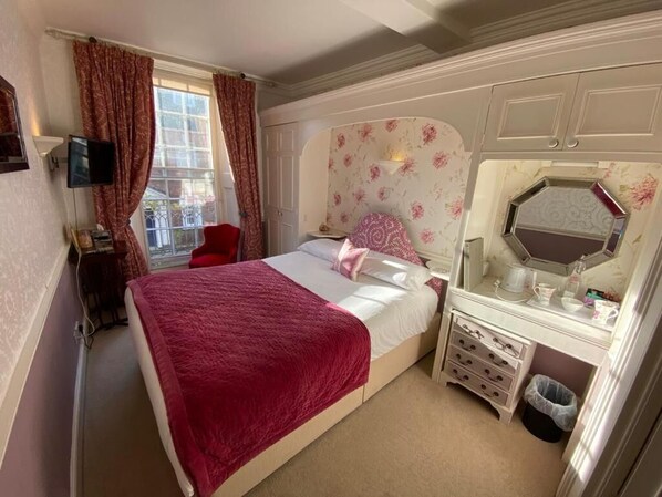 Un dormitorio con una cama, una tocador con espejo, una ventana con cortinas y un televisor en la pared.