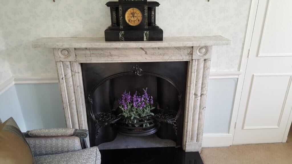 A fireplace with a clock above it, a flower arrangement in the hearth, and a chair to the left.