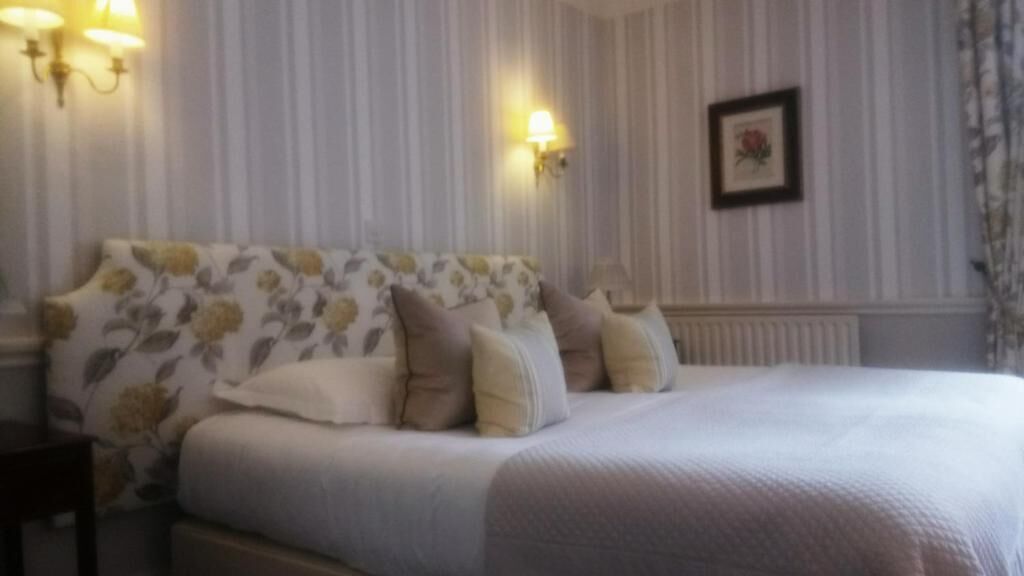 A hotel room with a bed, floral-patterned headboard, two wall-mounted lamps, and a framed picture on the wall.