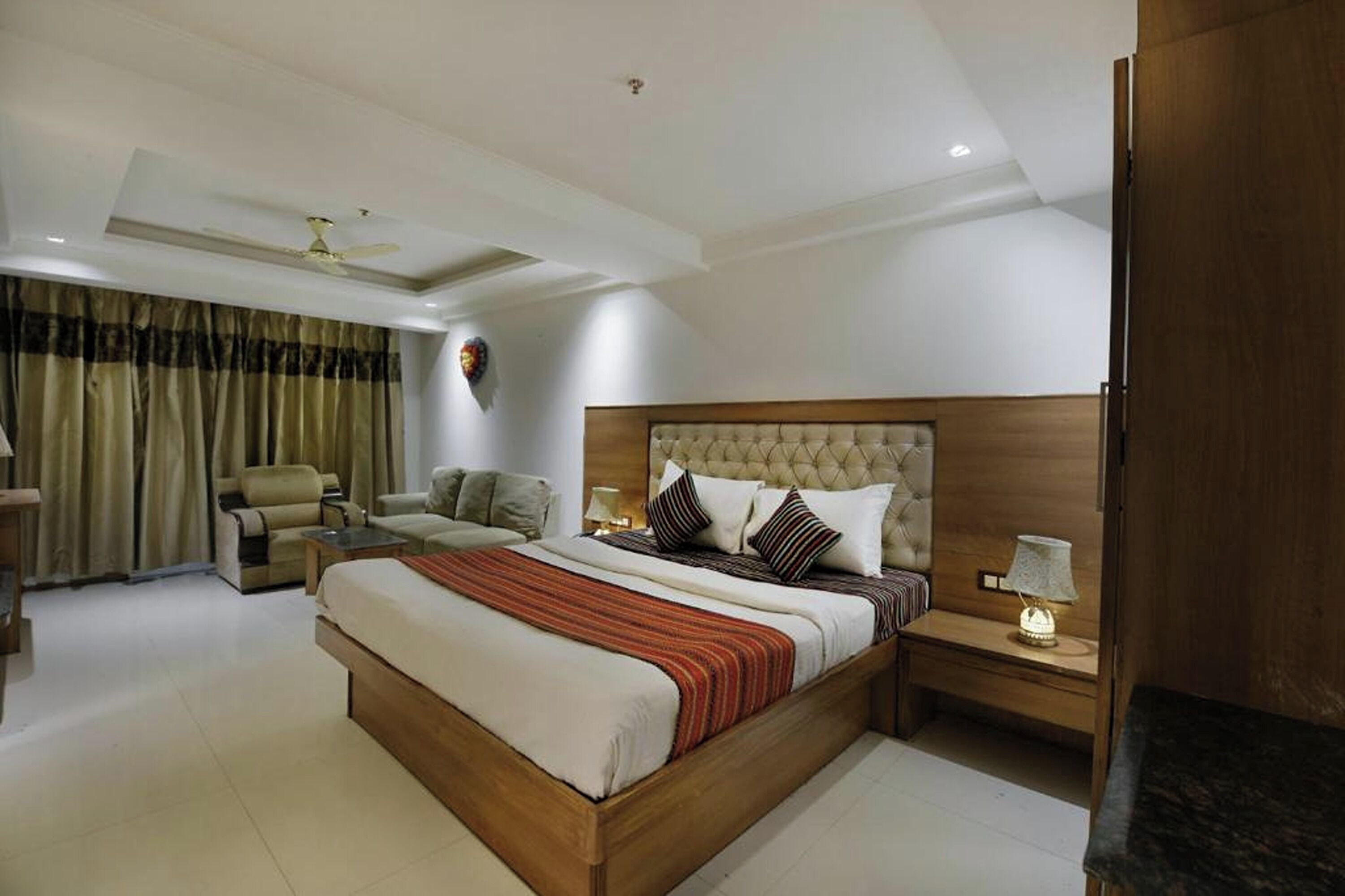 A modern bedroom with a large bed, a wooden headboard, a bedside table with a lamp, and a seating area with a chair and a sofa.