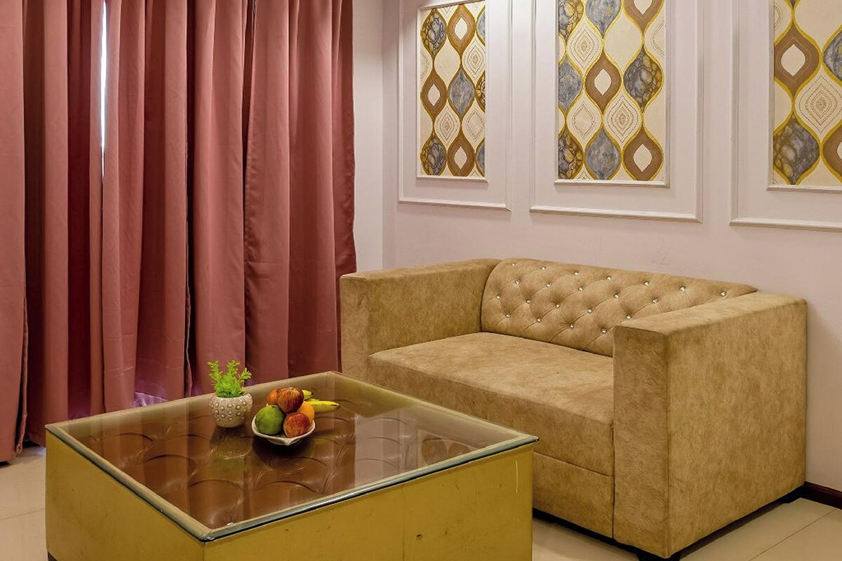 A living room with a beige sofa, a glass coffee table with fruits, a ceiling fan, and patterned wall art.