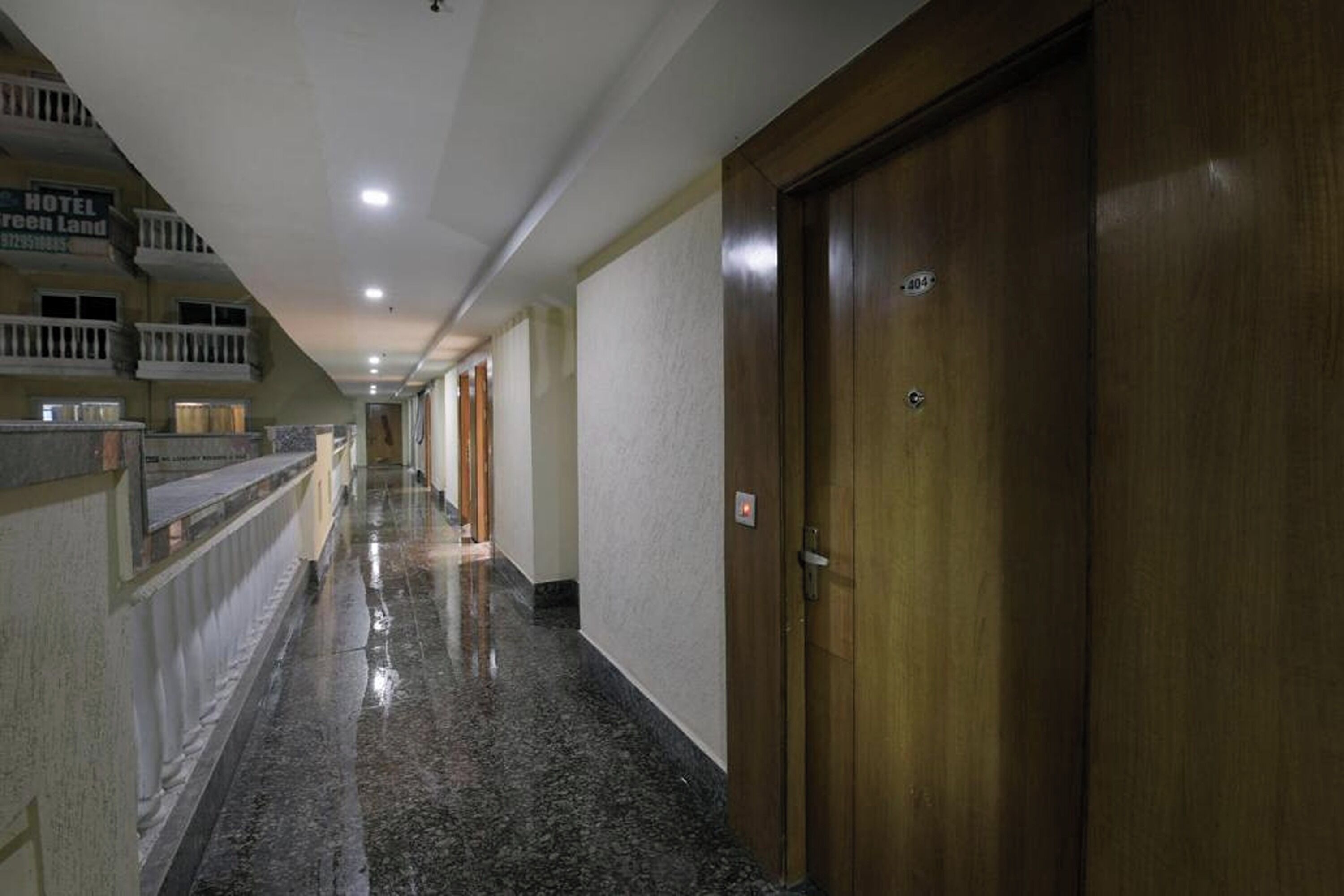 A hotel corridor with a wooden door on the right, a reception area with a balcony on the left, and a marble floor.